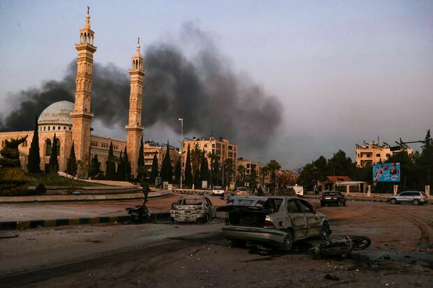 Columnas de humo a lo lejos mientras se ven automóviles dañados en el lugar de los ataques aéreos del régimen sirio contra combatientes antirrégimen en Alepo, en el norte de Siria, el 30 de noviembre de 2024. (Foto de AAREF WATAD / AFP)